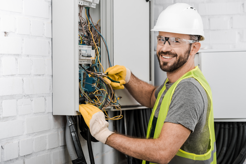 smiling-handsome-electrician-repairing-electrical-box-with-pliers-in-corridor-and-looking-at-camera